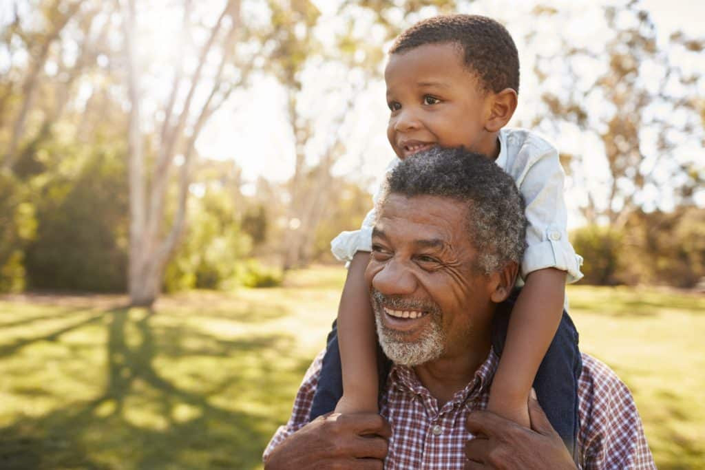 Grandfather Carries Grandson On Shoulders During Walk In Park
