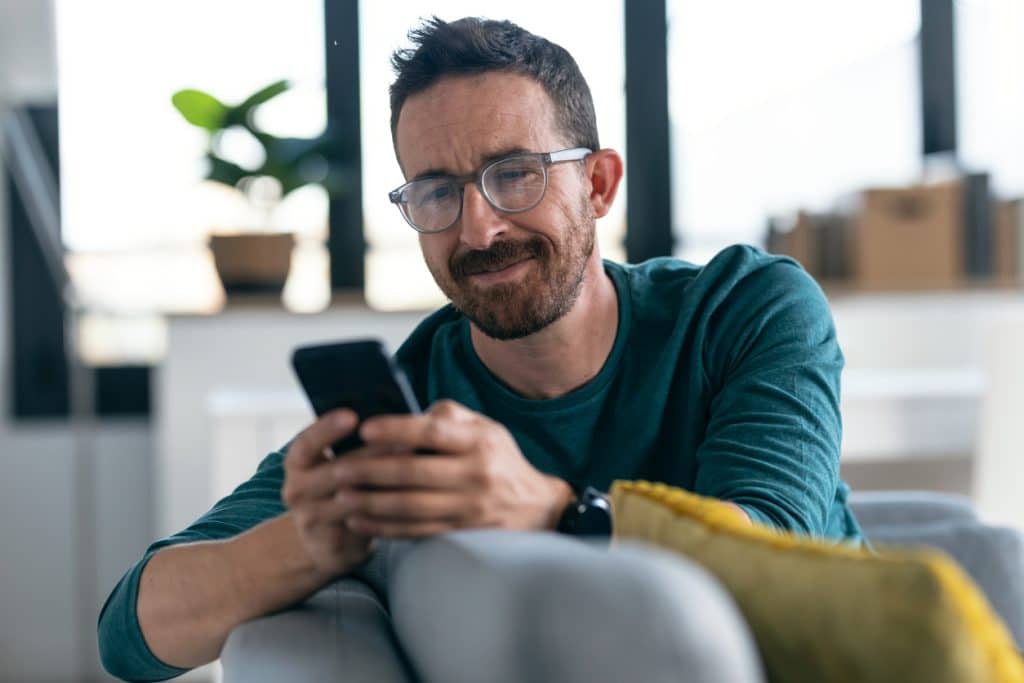 Mature happy man with eyeglasses using his smartphone while relaxing in the couch at home